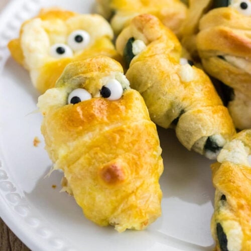 Closeup of jalapeno halves filled with cheese and wrapped in golden brown crescent roll dough to look like spooky mummies.