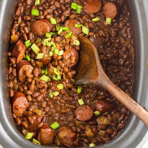 An overhead view of baked beans and kielbasa in the slow cooker with a wooden spoon.