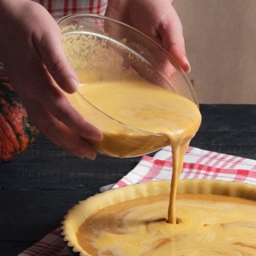 Closeup of hands pouring pumpkin pie filling into a pie crust.
