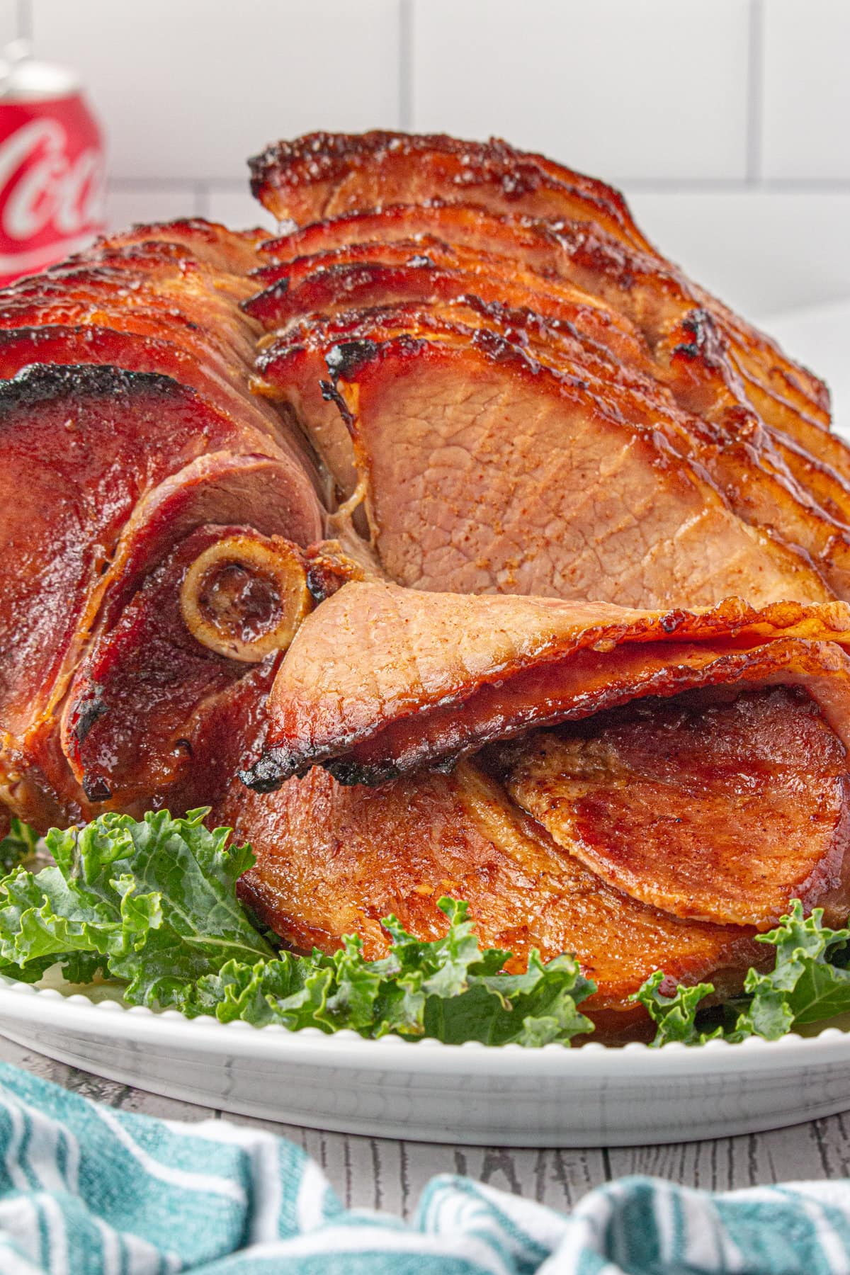 A spiral-cut ham, sliced with caramelized, glazed edges, sits on a platter and a coca-cola can is featured in the background.
