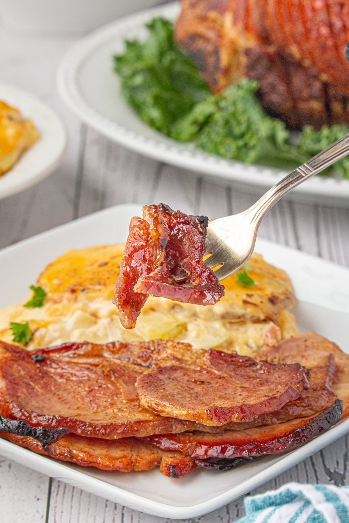 A fork holding a bite of Southern baked ham, hovering over a plate of ham with caramelized edges. Potatoes and more ham are in the background.