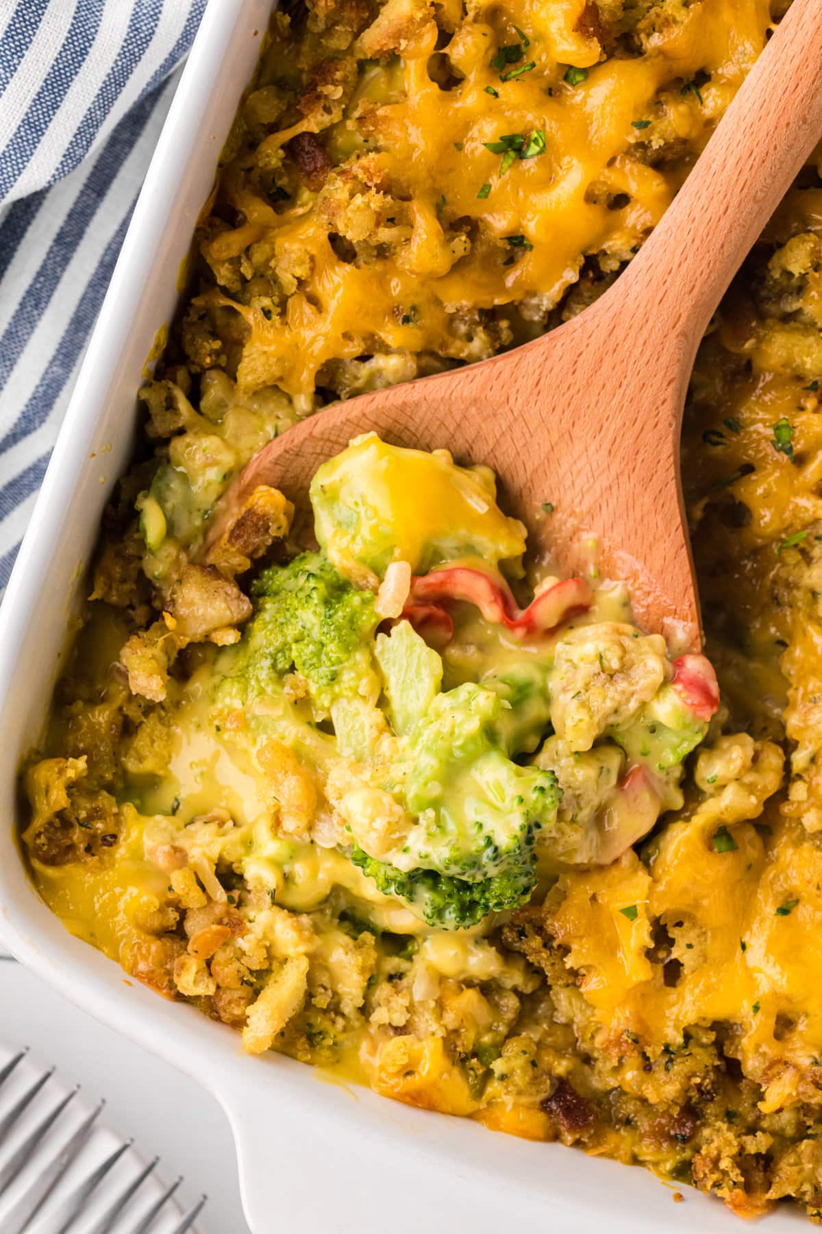 A wooden spoon scooping cheesy broccoli stuffing casserole out of a white baking dish.