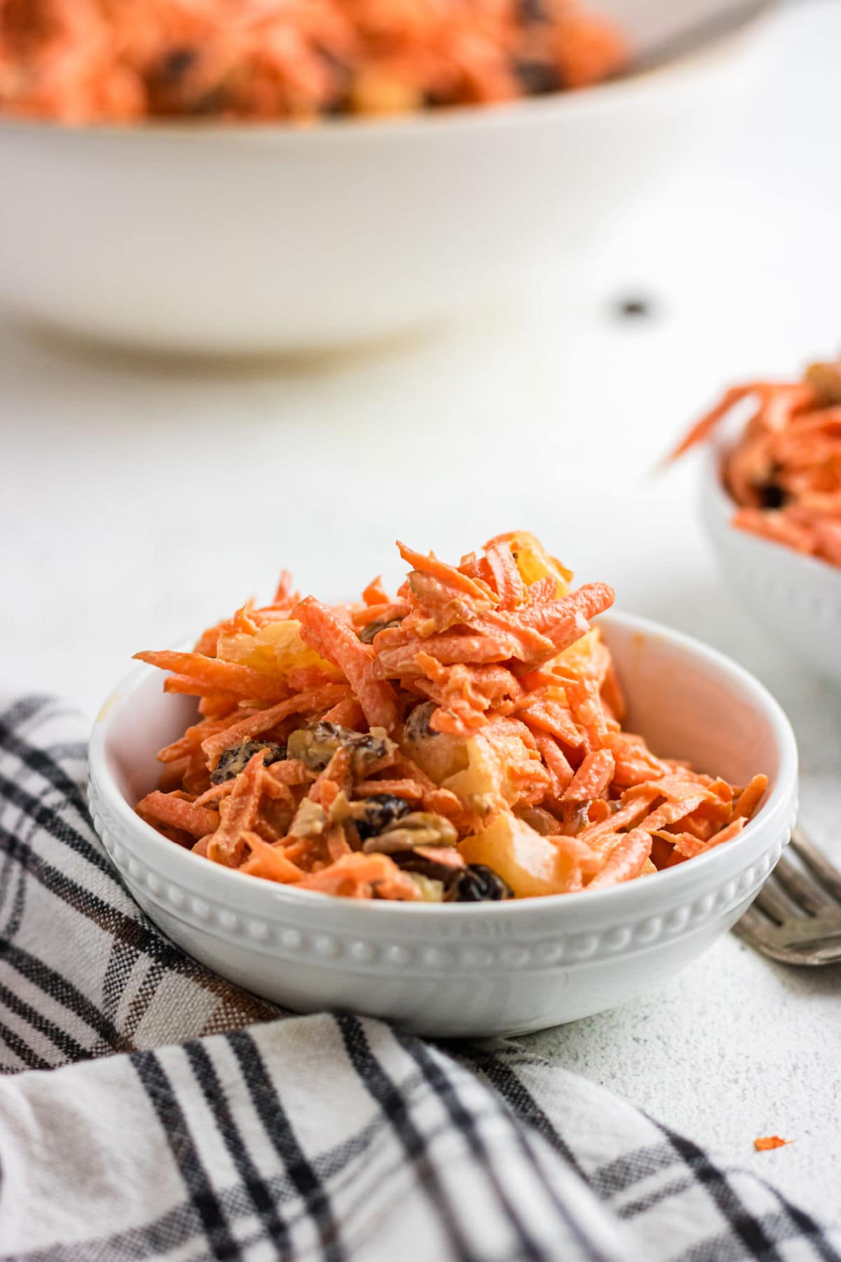 A serving of carrot raisin salad with pineapple and pecans in a small, white bowl.