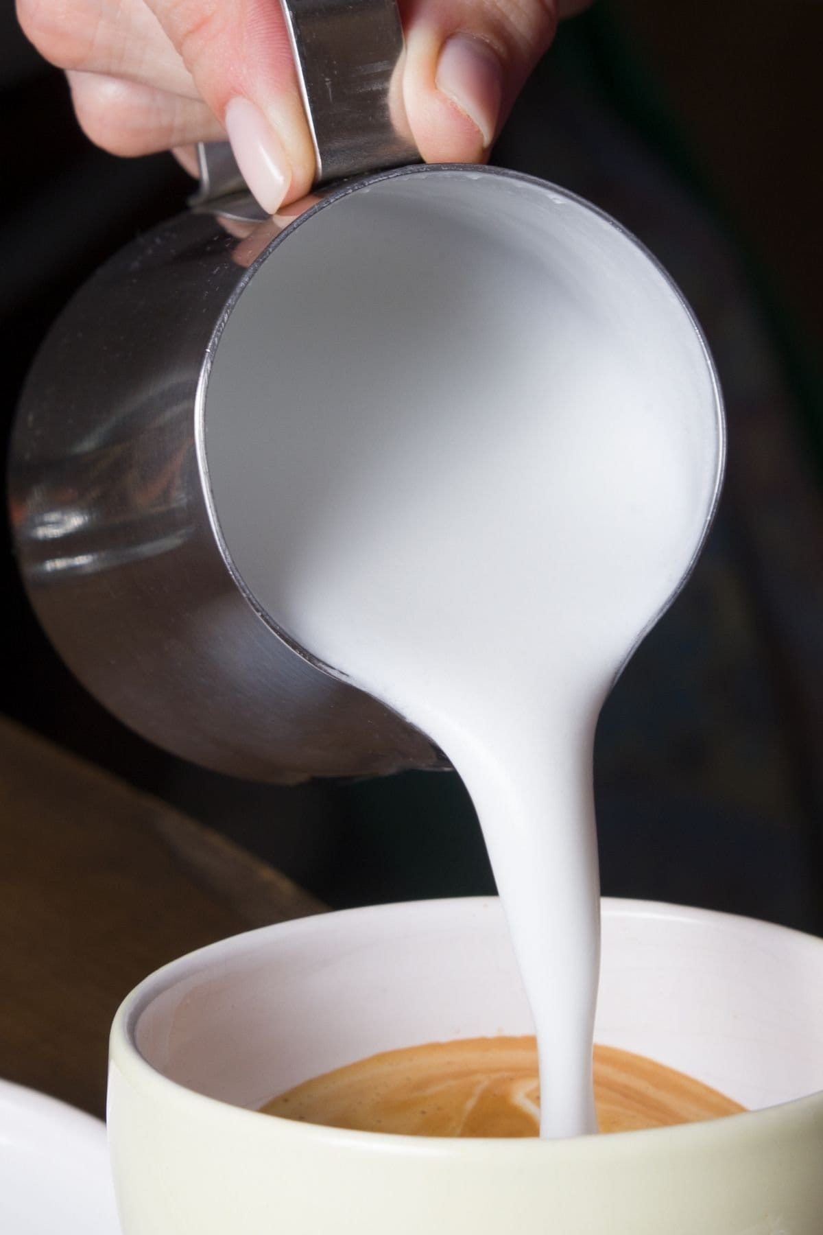 Thick, frothed milk pouring out of a cup onto a hot cup of coffee.