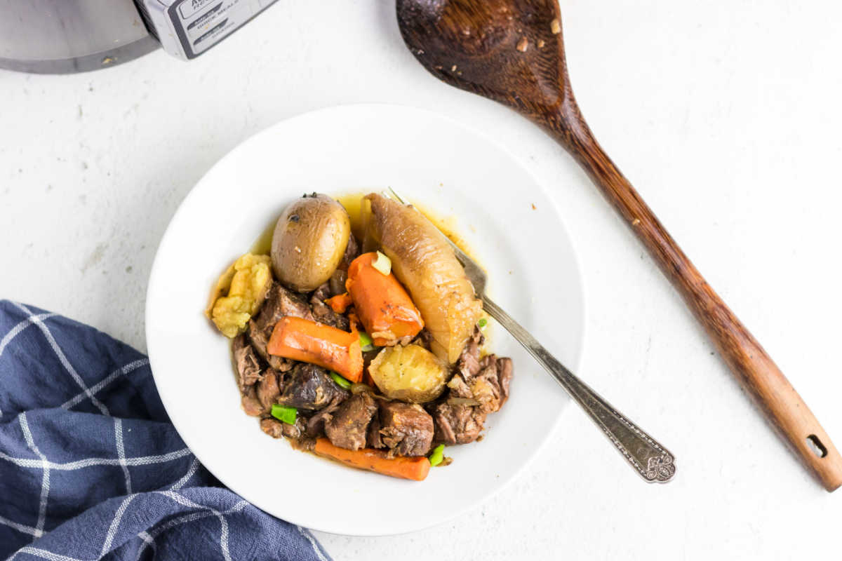 Beef stew with beef, potatoes, and carrots in a shallow bowl with a fork and wooden spoon next to it.