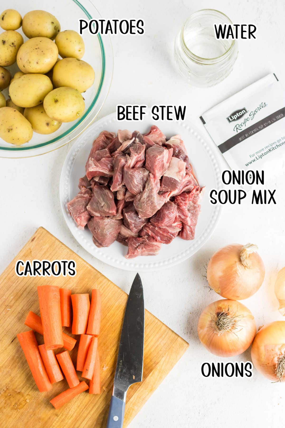 An overhead photo of stew ingredients: carrots on a cutting board; beef stew pieces on a plate; potatoes in a bowl; and onions, onion soup mix, and water off to the side.