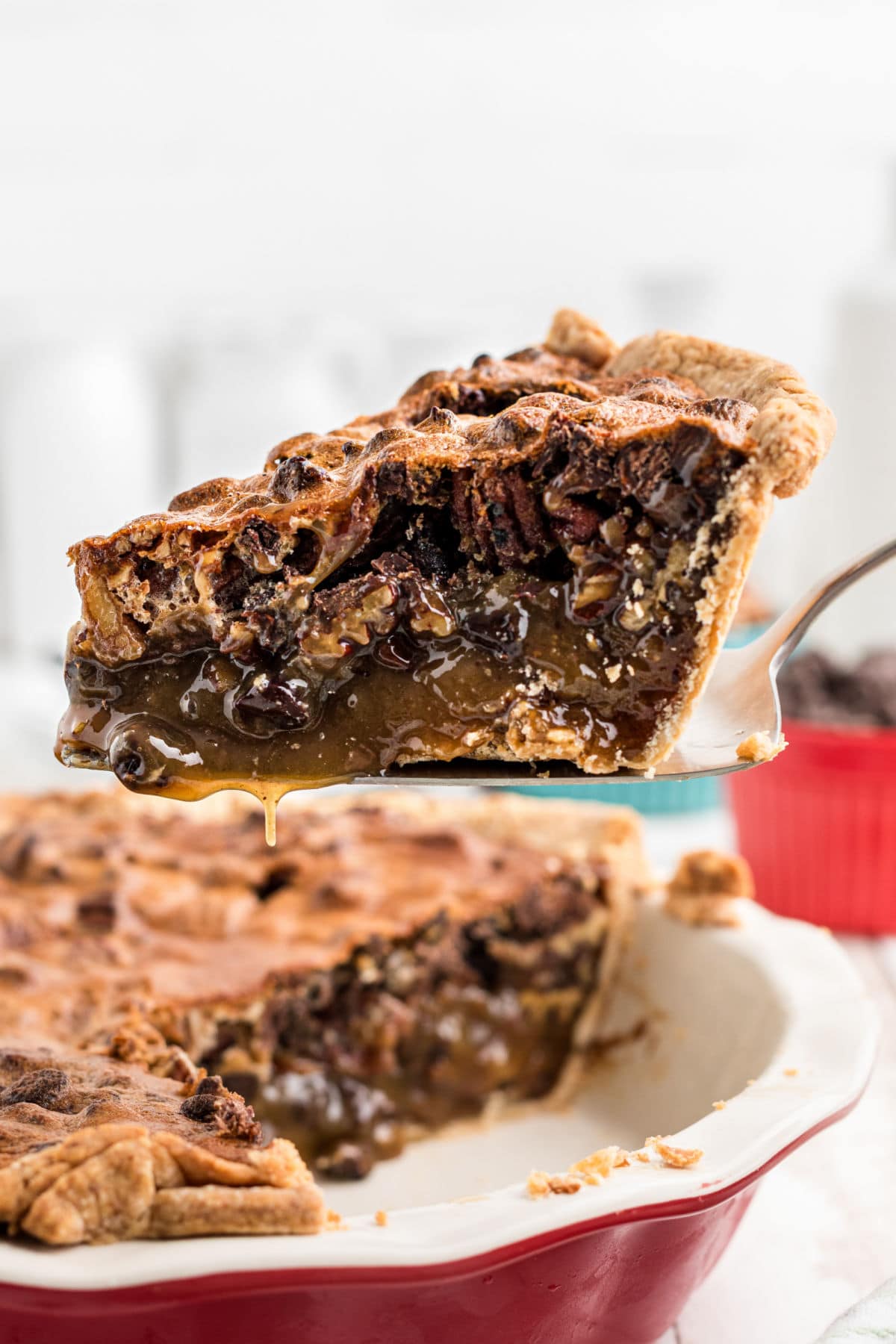 A slice of chocolate bourbon pecan pie on a pie server hovering over the rest of the pie in a red pie dish.
