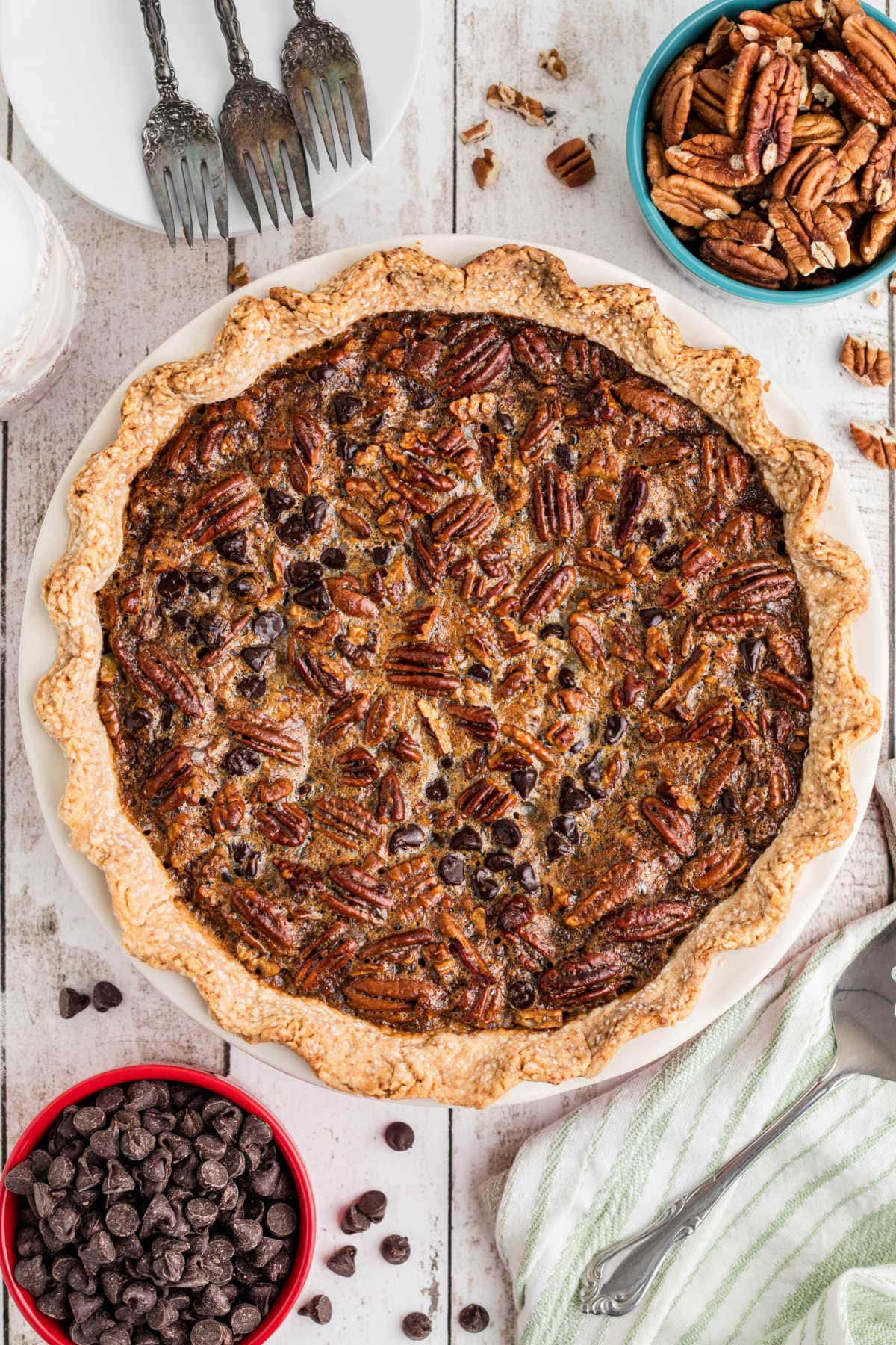 An overhead photo of a fully baked Chocolate Bourbon Pecan Pie with pecans, chocolate chips, and forks scattered around the frame of the photo.