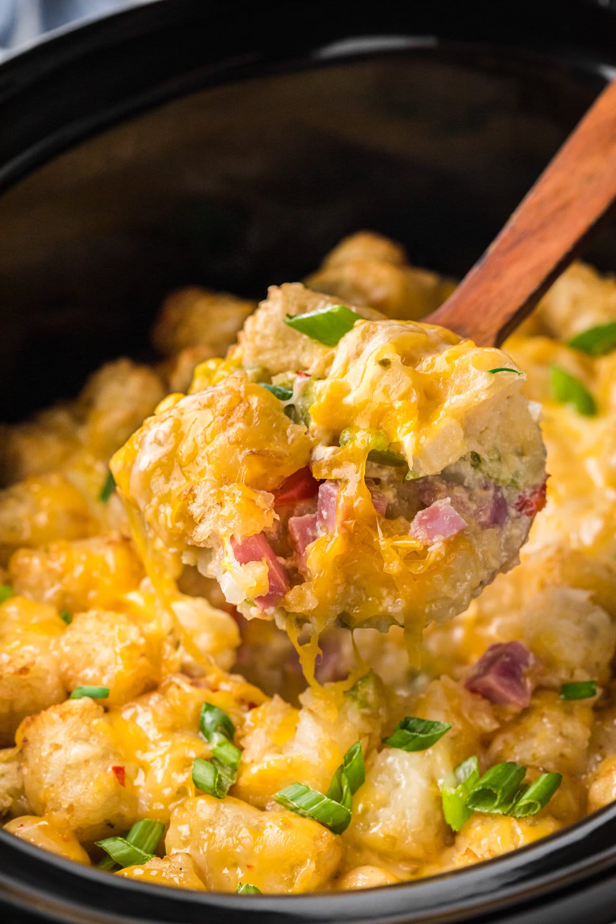 A close-up photo of tater tot casserole in a crockpot. A scoop of cheesy potatoes, ham, and peppers is in the foreground of the photo. 