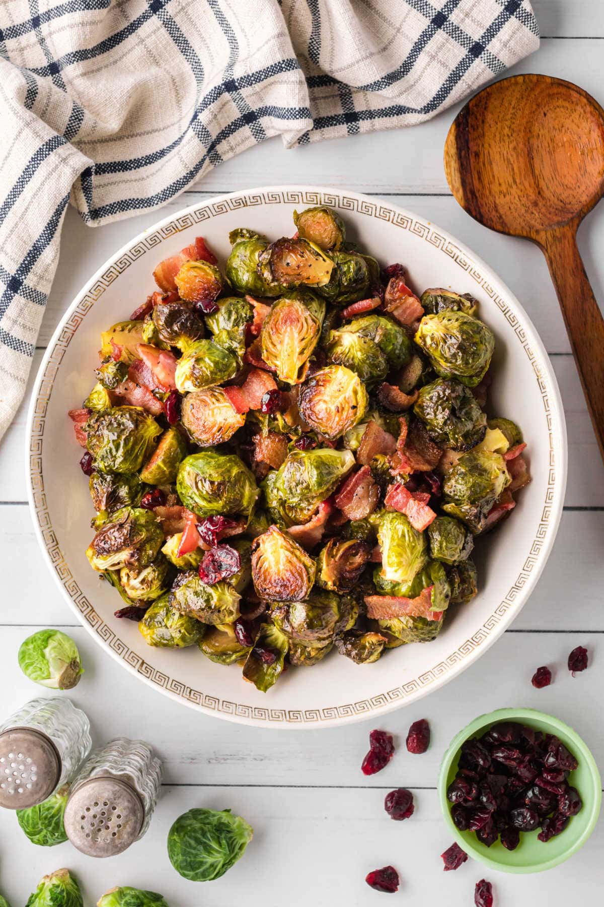 Crispy bacon and Brussels sprouts are shown in a white bowl with a big serving spoon next to it. There are cranberries tossed in with the veggies and some scattered around the outside of the bowl.