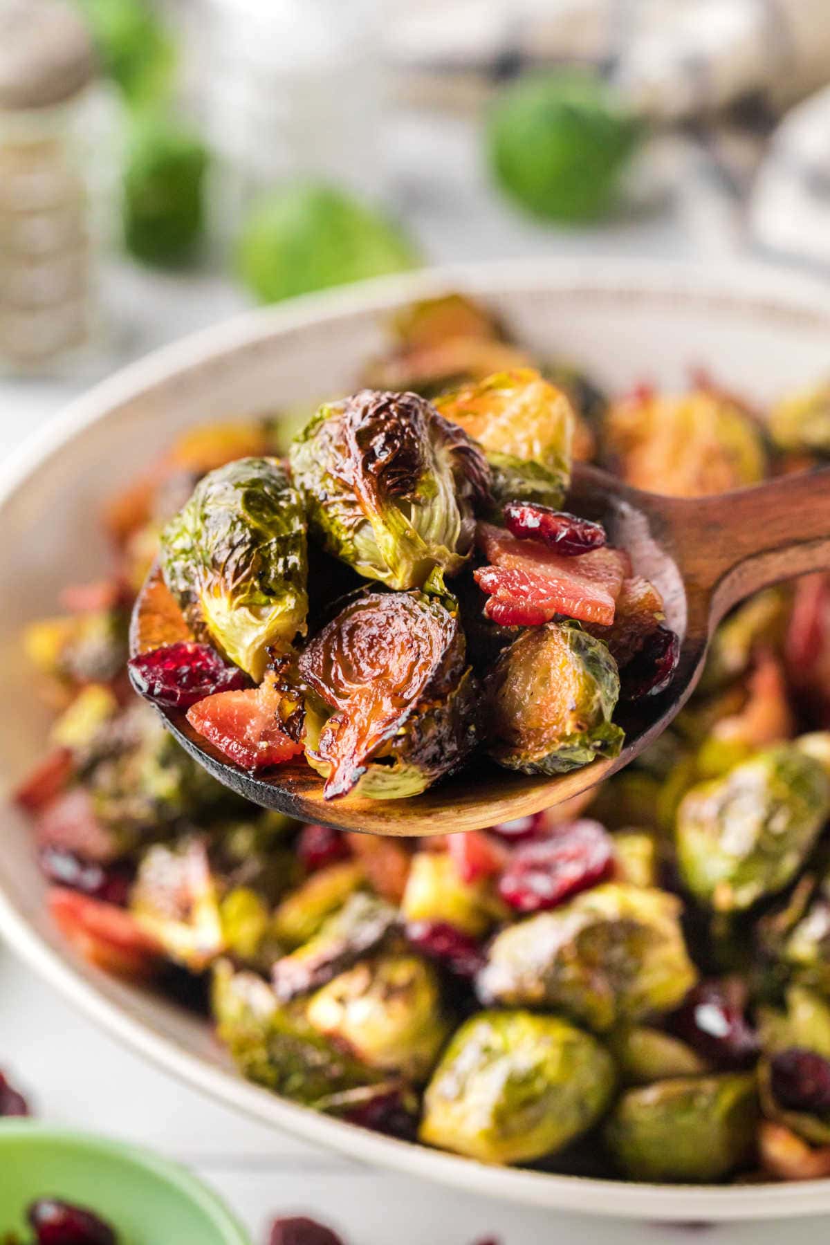 In the background, a large bowl with roasted bacon Brussels sprouts is shown. In the foreground, a large wooden spoon shows the camera a scoop of crispy Brussels sprouts, pieces of bacon, and cranberries.