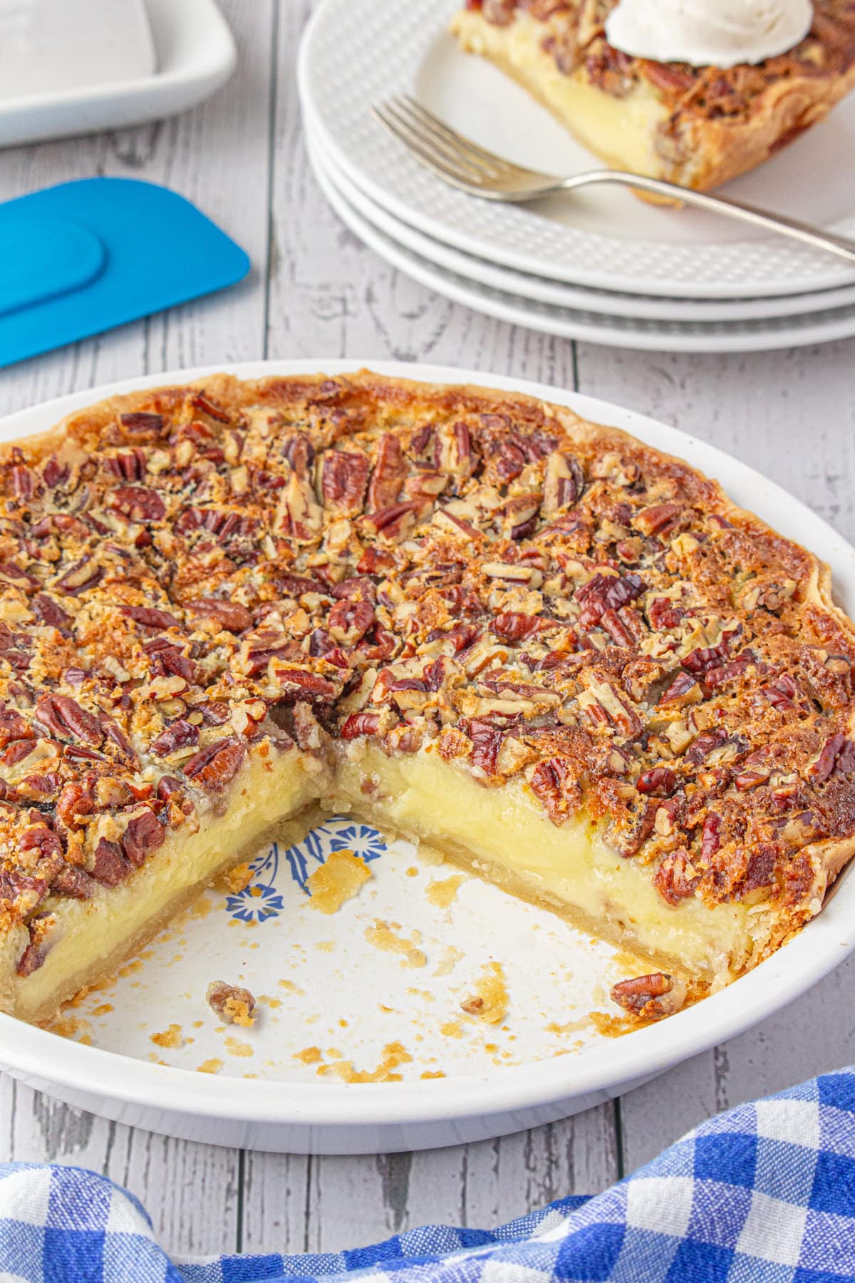 A buttermilk pecan pie with a large slice removed to a plate in the background. The interior of the pie shows pie crust topped with a buttermilk layer which is topped with pecans.