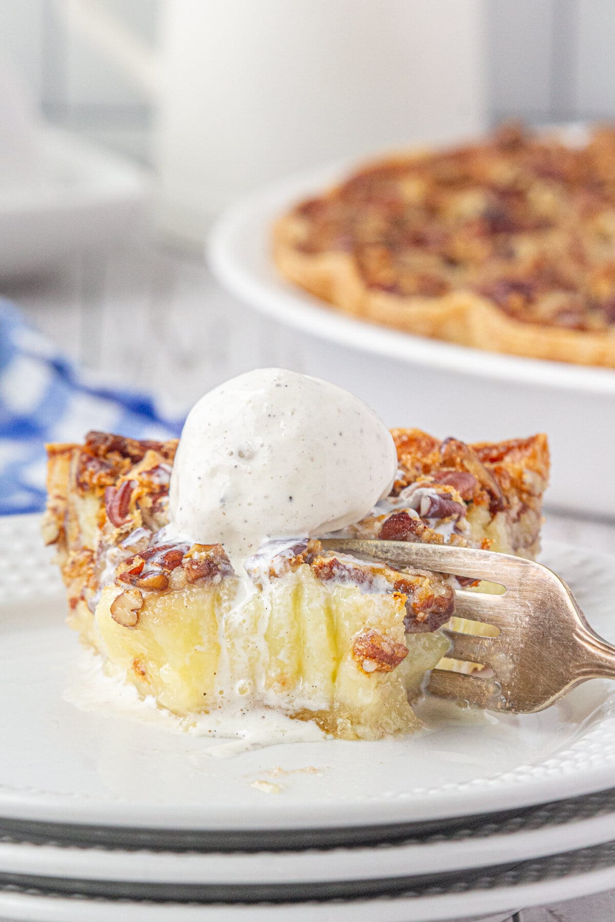 A close-up photo of a slice of buttermilk pie topped with ice cream. One bite is removed and a fork is shown slicing into the piece. 