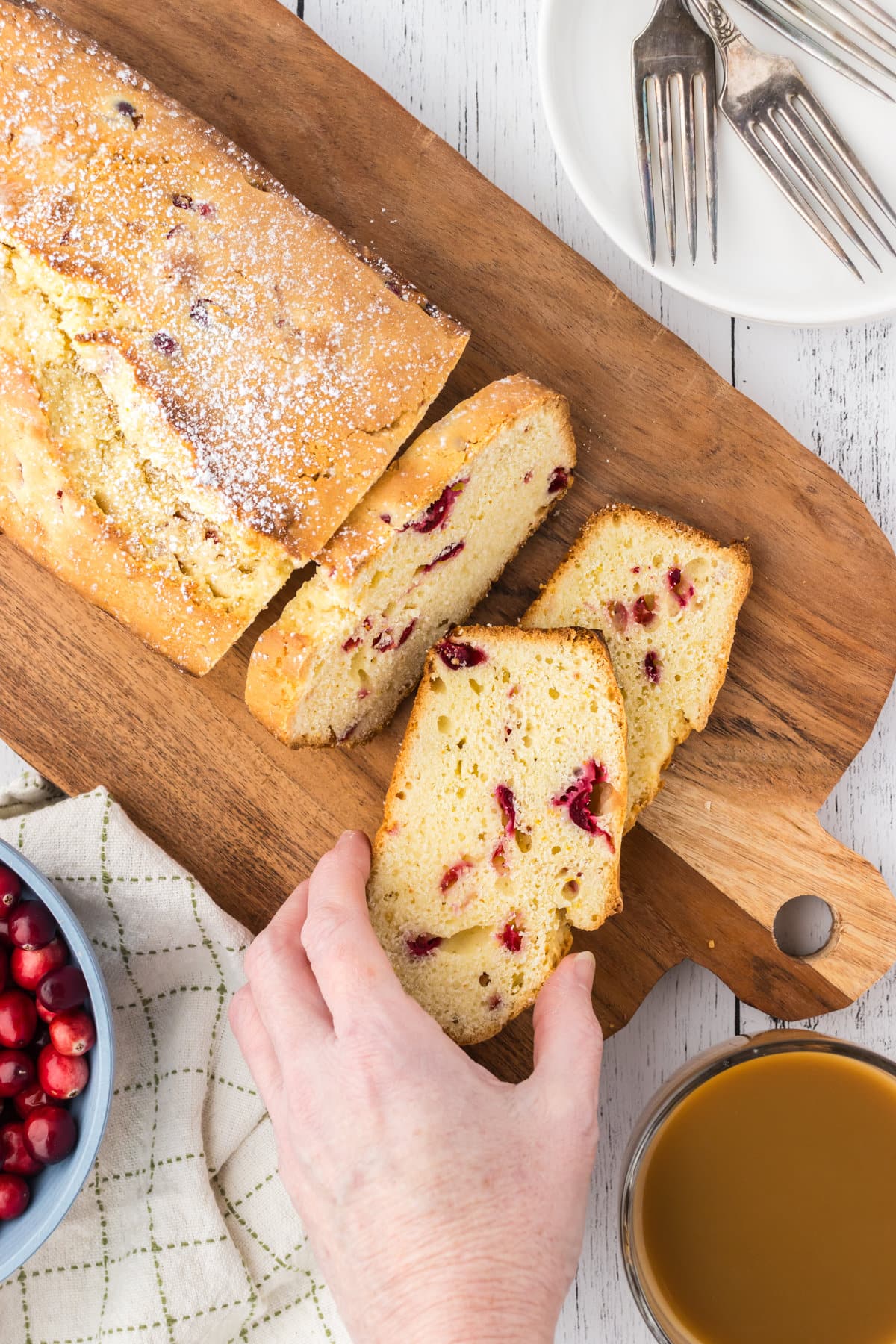 An overhead view of cranberry pound cake, sliced into a few pieces. A hand is holding one slice to show the cranberries inside the loaf.
