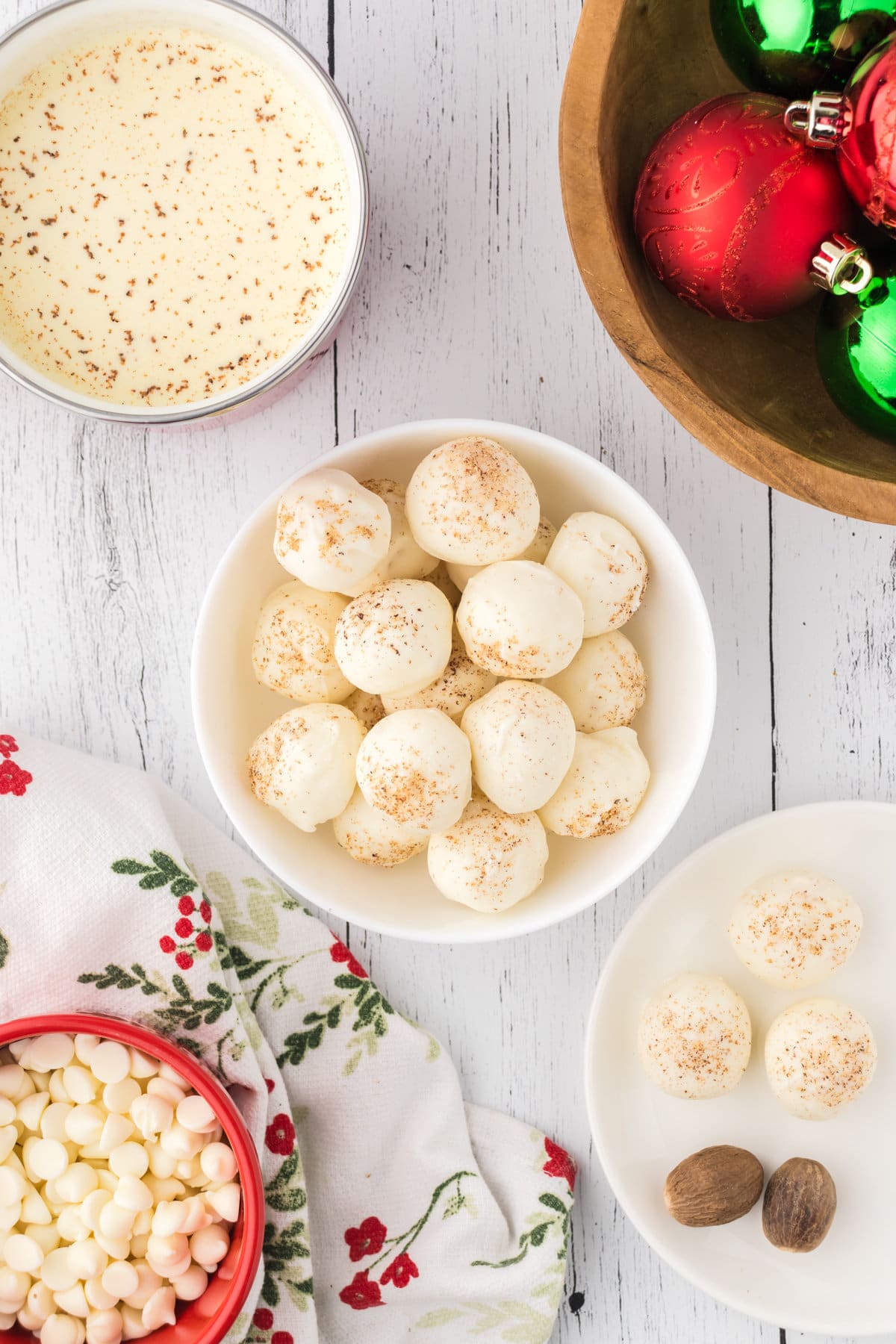 Eggnog truffles, sprinkled with nutmeg, in a white bowl. There are white chocolate chips, whole nutmegs, eggnog, and Christmas ornaments around the border.