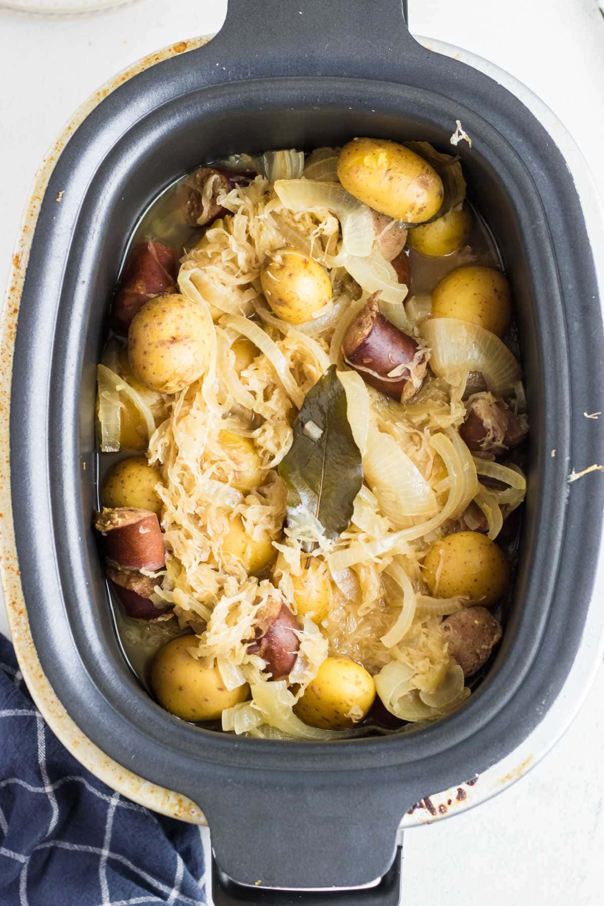 A crockpot filled with sliced kielbasa, potatoes, onions, and sauerkraut. A bay leaf rests on top.