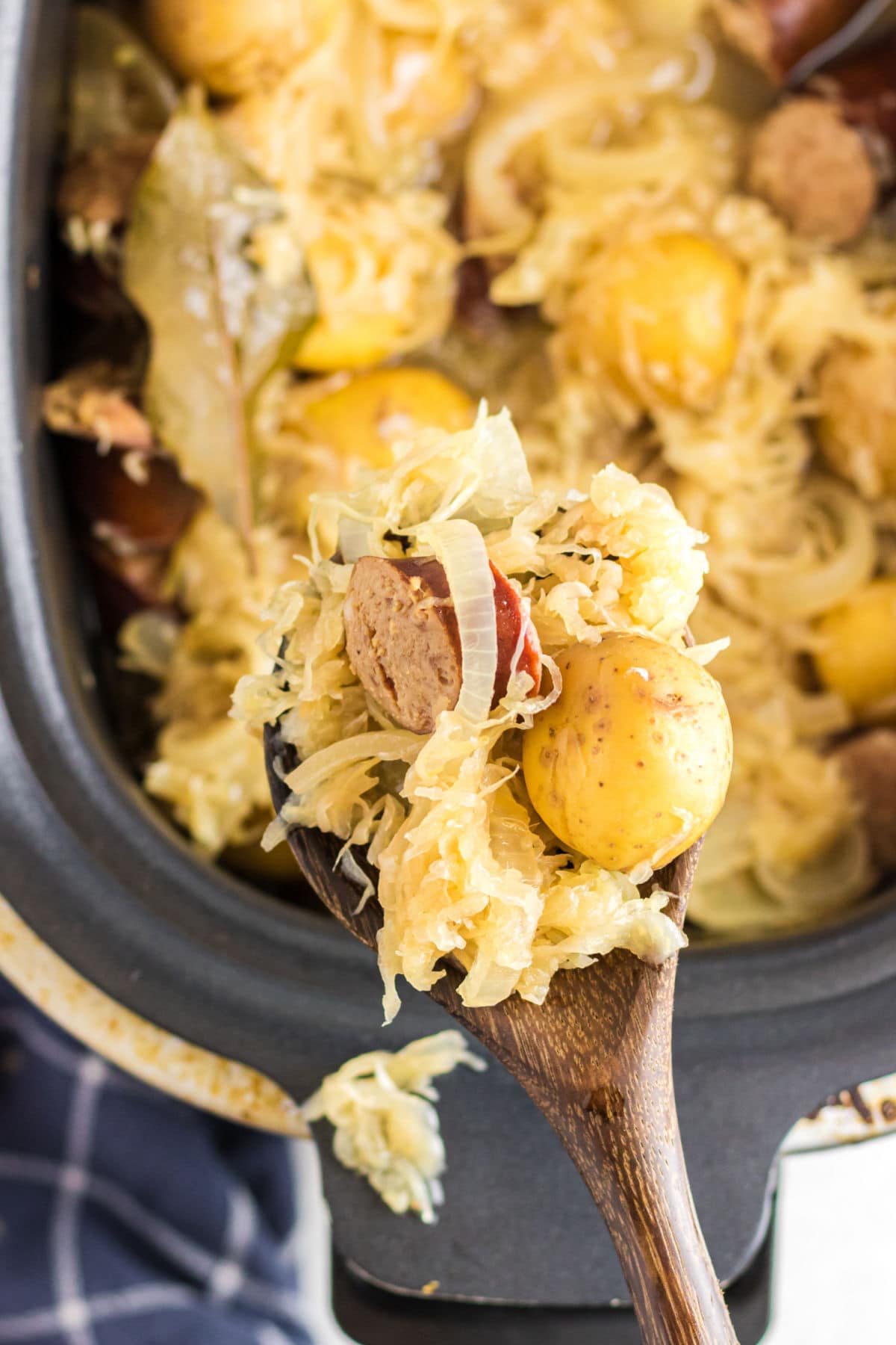 An overhead images of a scoop of kielbasa, sauerkraut, and potatoes on a wooden spoon hovers over a crockpot containing the rest of the food.