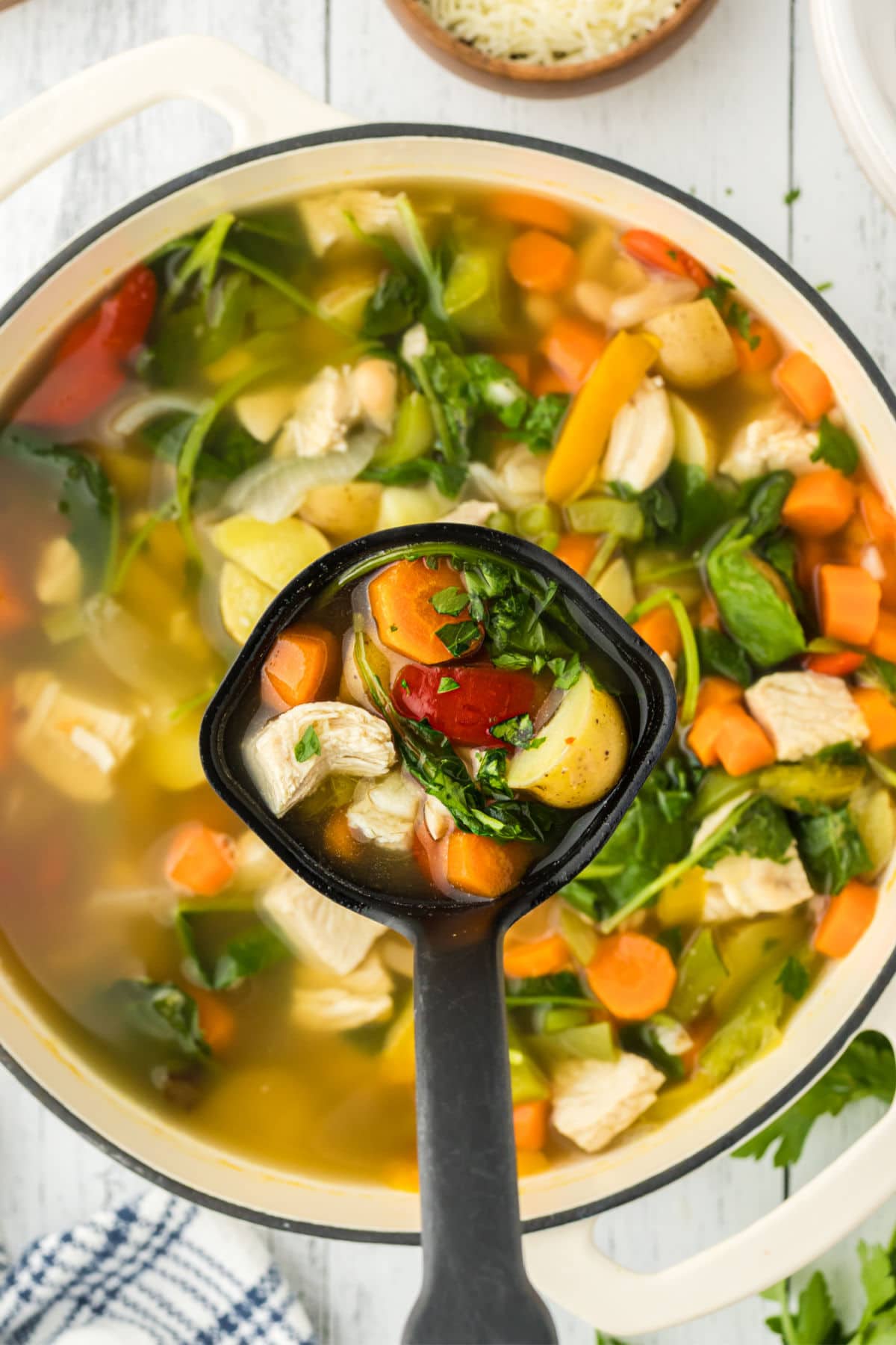 In the foreground, a ladle is shown scooping some Tuscan turkey soup, highlighting veggies, potatoes, and turkey in a broth. In the background is the rest of the soup in a white Dutch oven. 