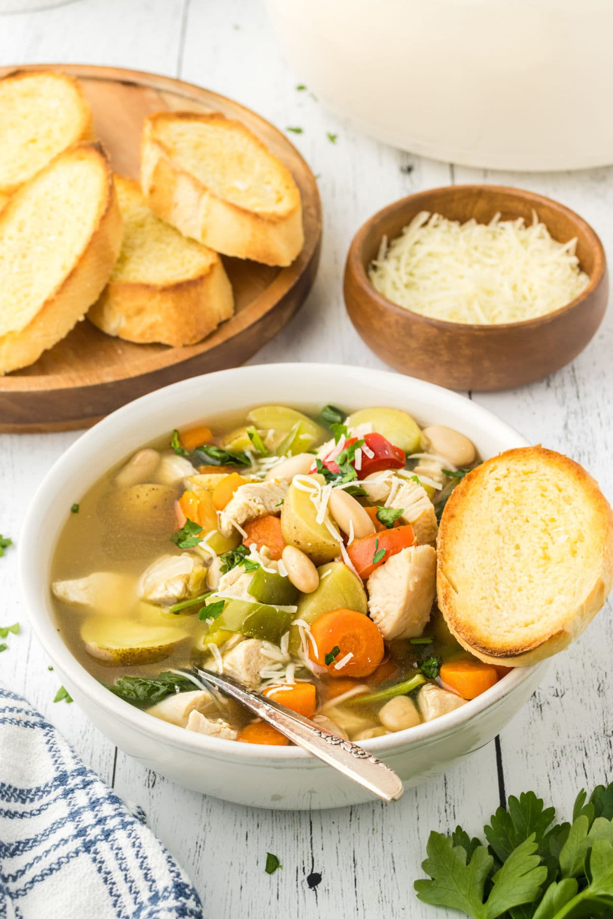 A bean, vegetable, potato, turkey, and broth-filled bowl sits at an angle to the camera. A toasted baguette slice is perched on the edge, and parmesan cheese is situated behind the soup bowl.