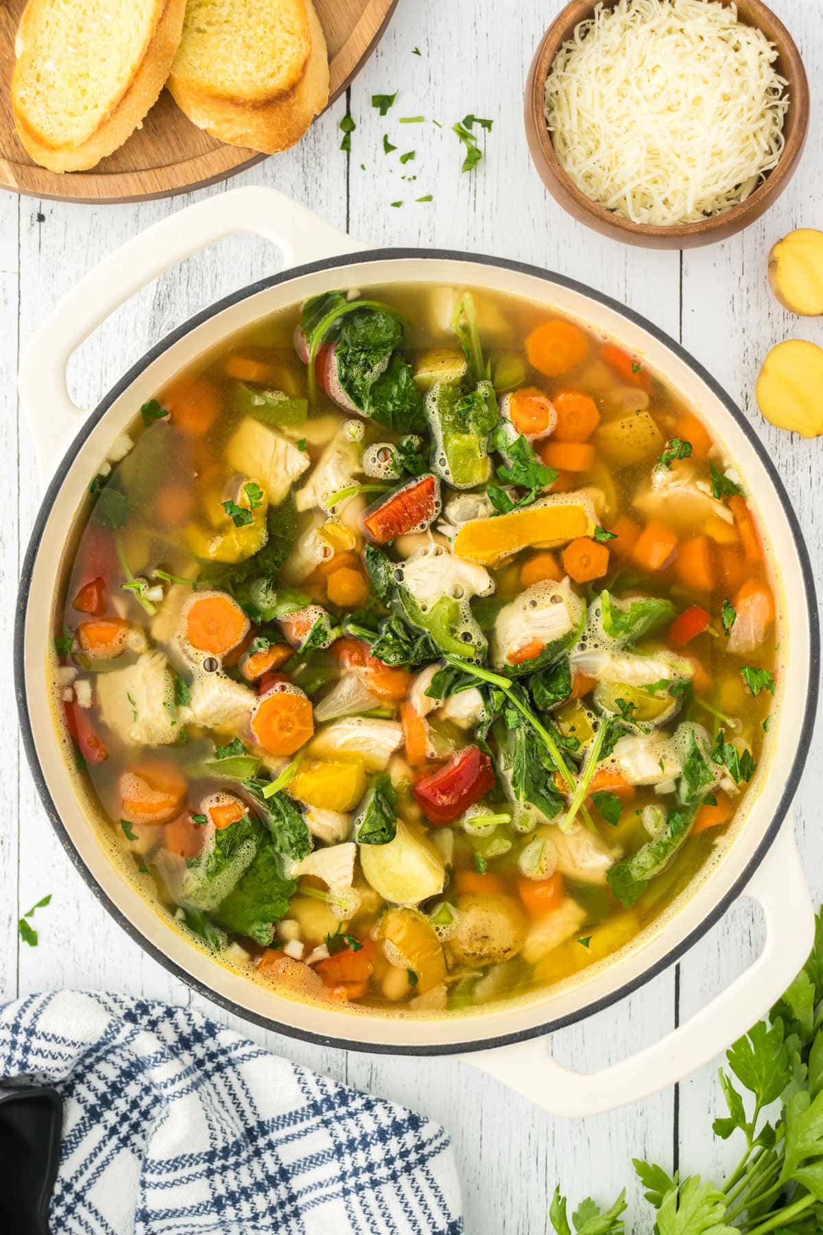 An overhead image of Tuscan turkey soup, showing pieces of kale, potatoes, carrots, peppers, beans, and broth in a white Dutch oven. Bread and parmesan cheese are off to the side of the image.