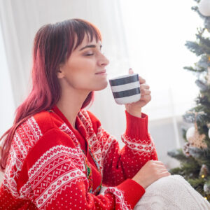A woman in a red sweater enjoying a cup of coffee next to a Christmas tree.