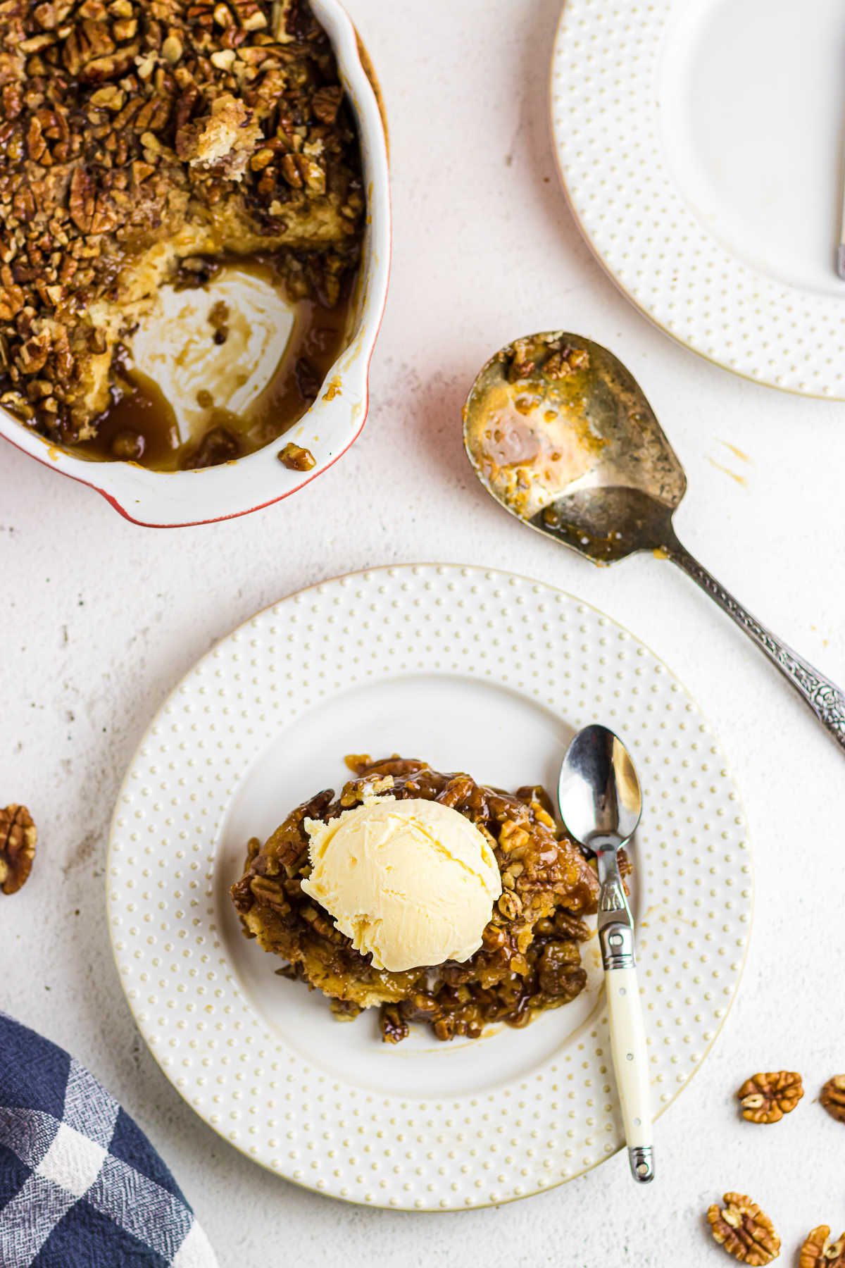 An overhead photo of pecan pie cobbler topped with ice cream on a white plate. Gooey pecans are shown leaking out from the sides of the scoop, and the casserole dish with the remaining cobbler is shown off to the corner of the photo.