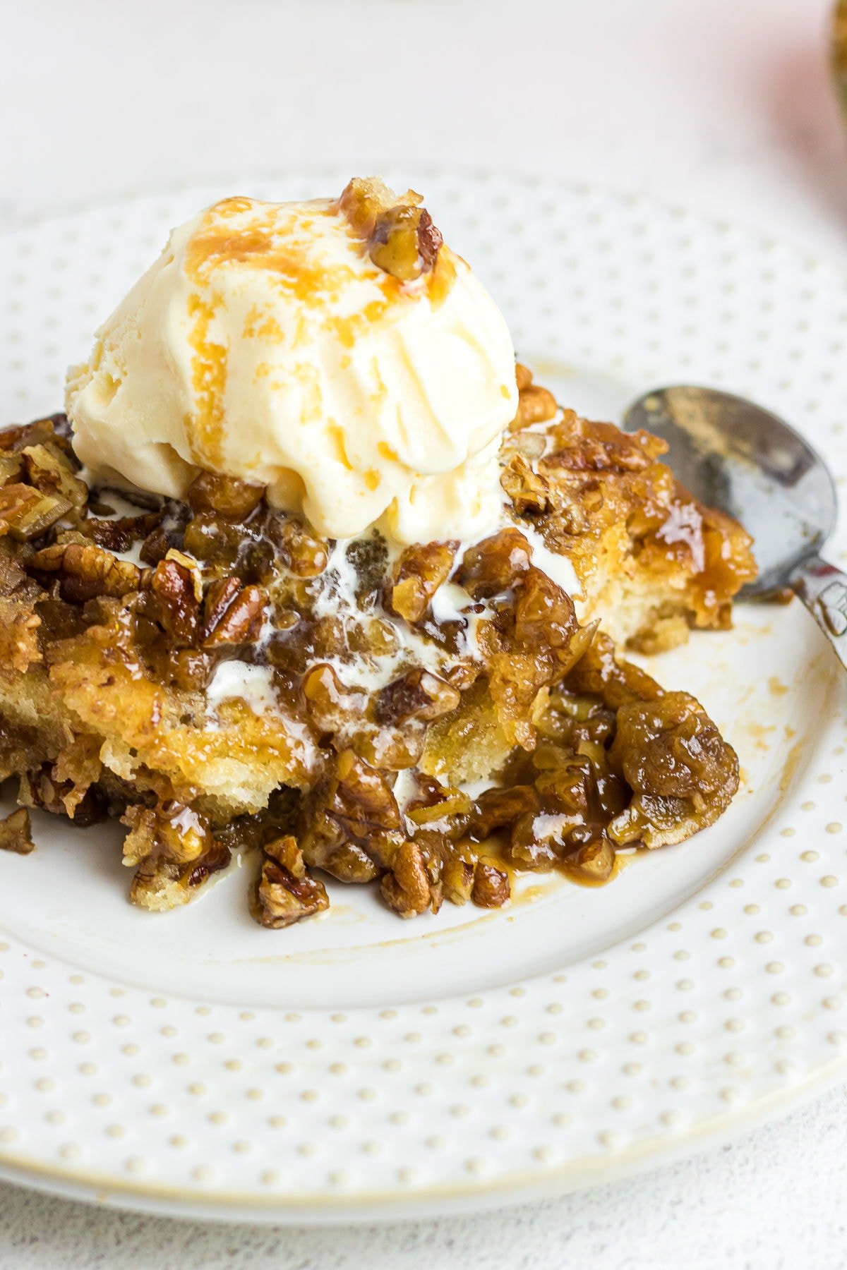 A close-up photo highlights a scoop of gooey pecan cobbler on a white plate with some ice cream on top and a spoon in the background.