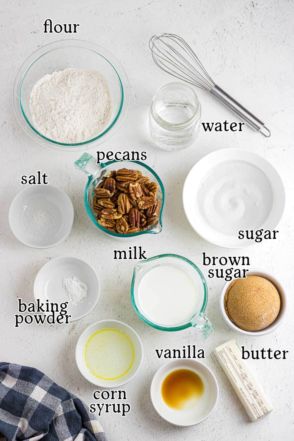 An overhead shot with text overlay showing the raw ingredients for pecan pie cobbler, including flour, pecans, sugar, milk, vanilla, butter, salt, baking powder, and water in small bowls.