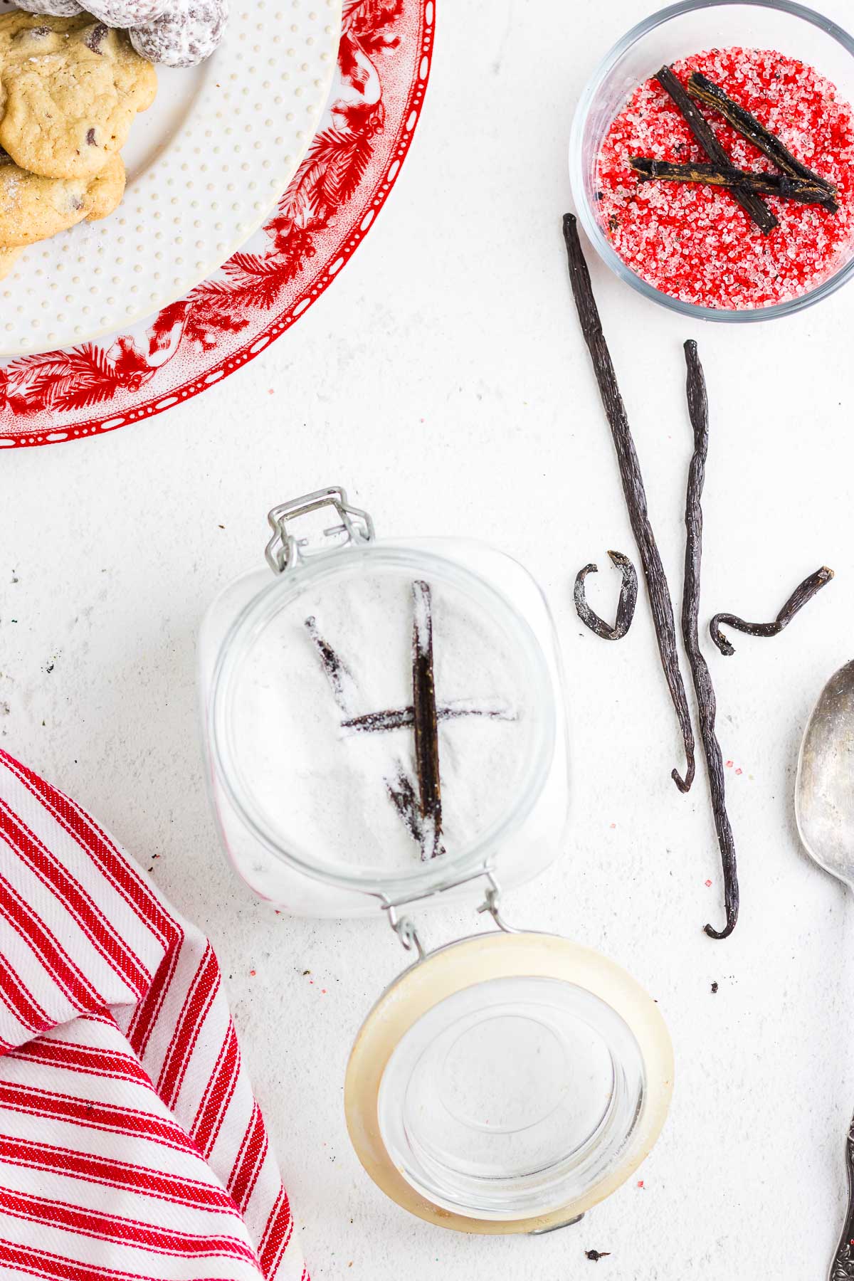 A lidded mason jar is open to reveal white vanilla sugar with vanilla bean pods inside. There is a small bowl off to the side with red granulated vanilla sugar and vanilla bean pods.