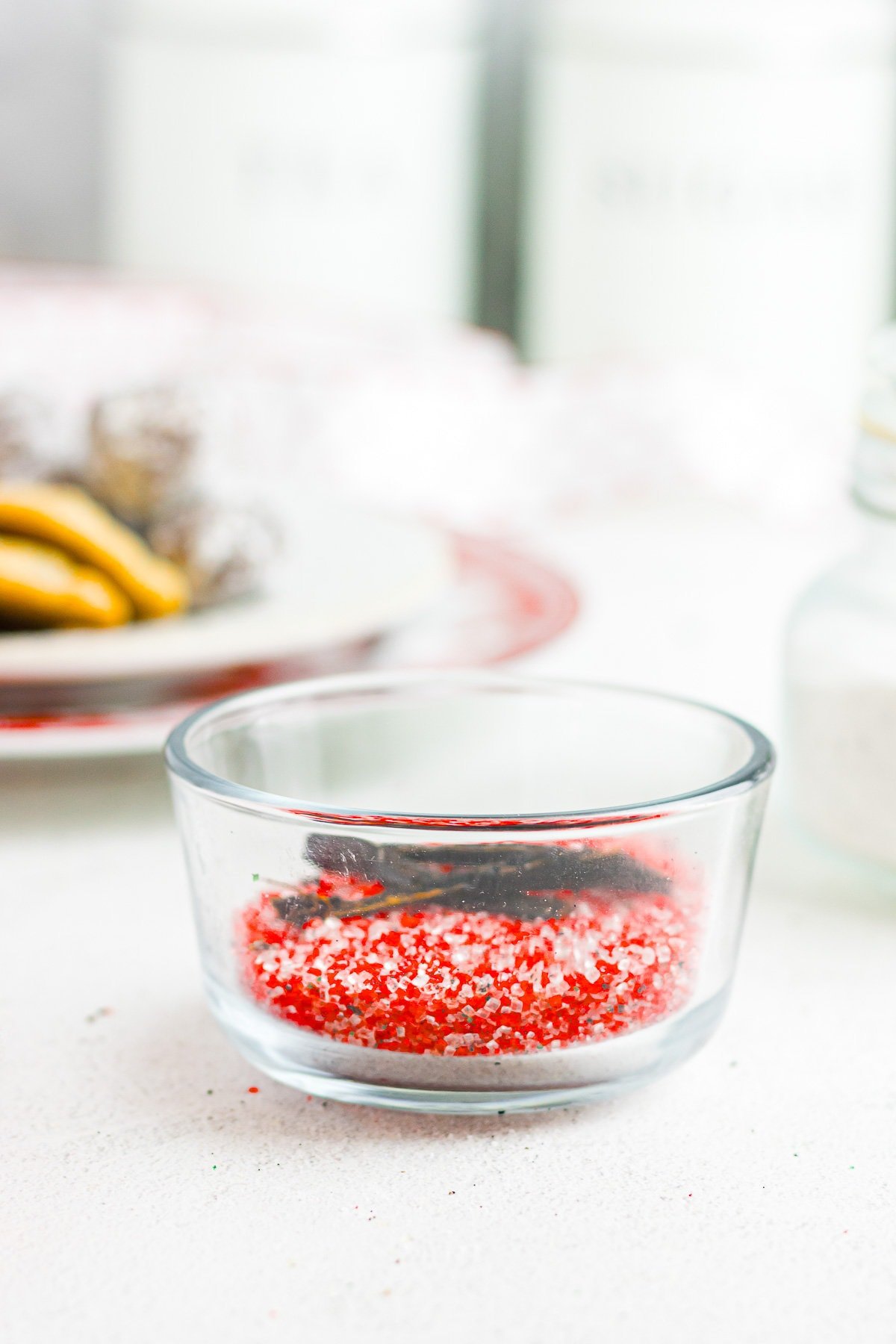 A glass bowl filled with vanilla sugar made with red sugar crystals. Vanilla bean pods lay on top of the sugar.