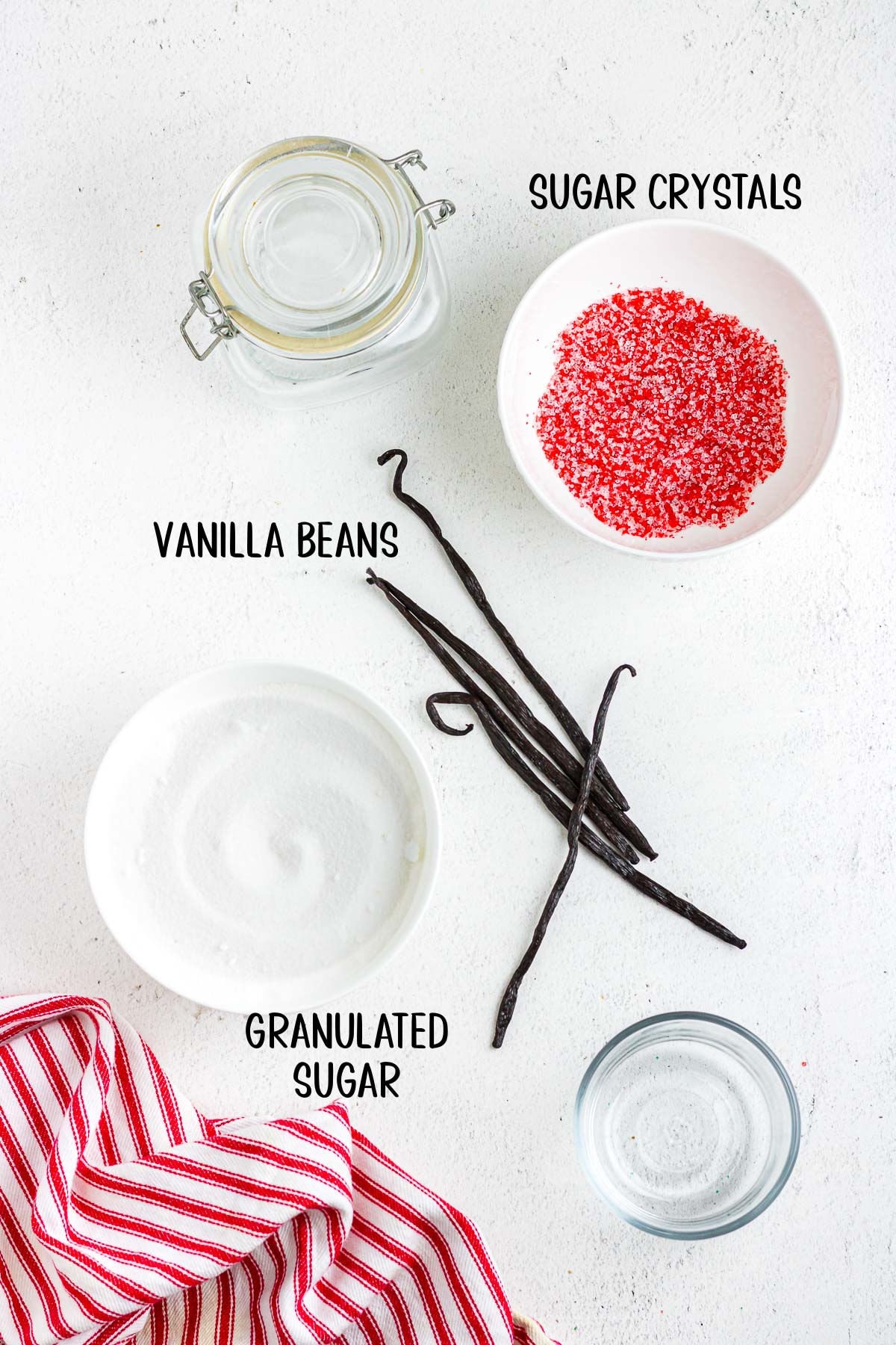 A bowl of granulated sugar, vanilla beans, and a bowl of red sugar crystals sit next to a mason jar in this overhead photo with text overlay.