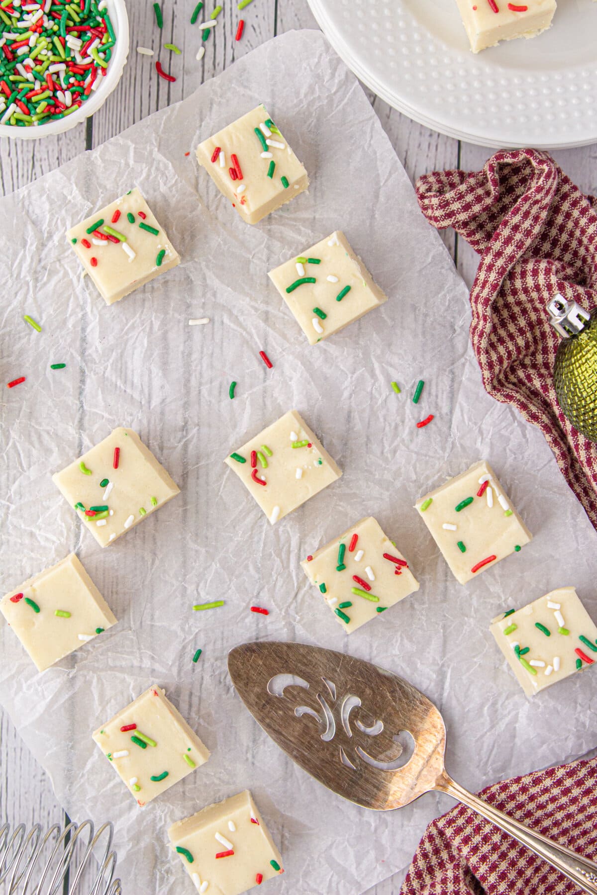 Pieces of white fudge are topped with Christmas-colored sprinkles and set on a piece of parchment paper.