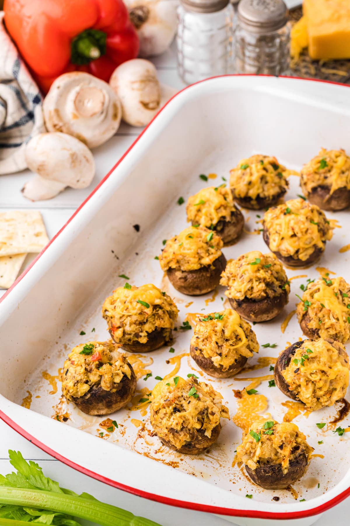 Cheese-covered crab-stuffed mushrooms in a baking dish, photographed at an angle. Extra ingredients are shown in the background.