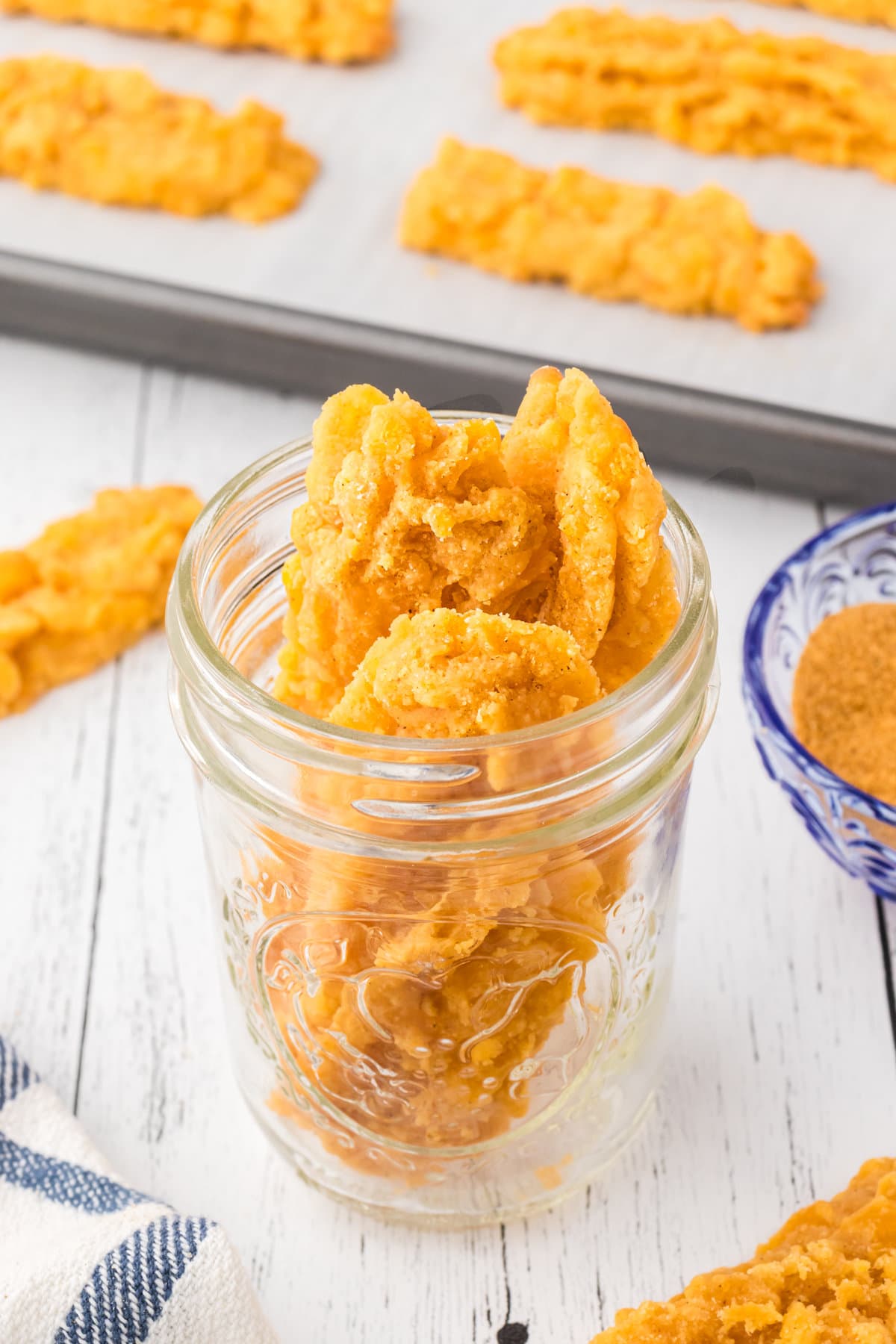 Crispy cheese straws in a glass jar and leftovers on a baking sheet in the background.