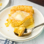 A square of fluffy spoon bread on a white plate with butter melting on top.