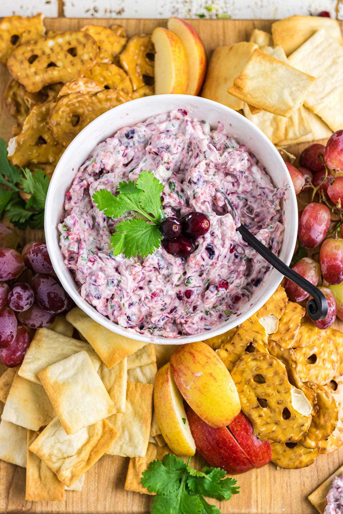 Cranberry jalapeno dip in a white bowl surrounded by chips, pretzels, and fruit.
