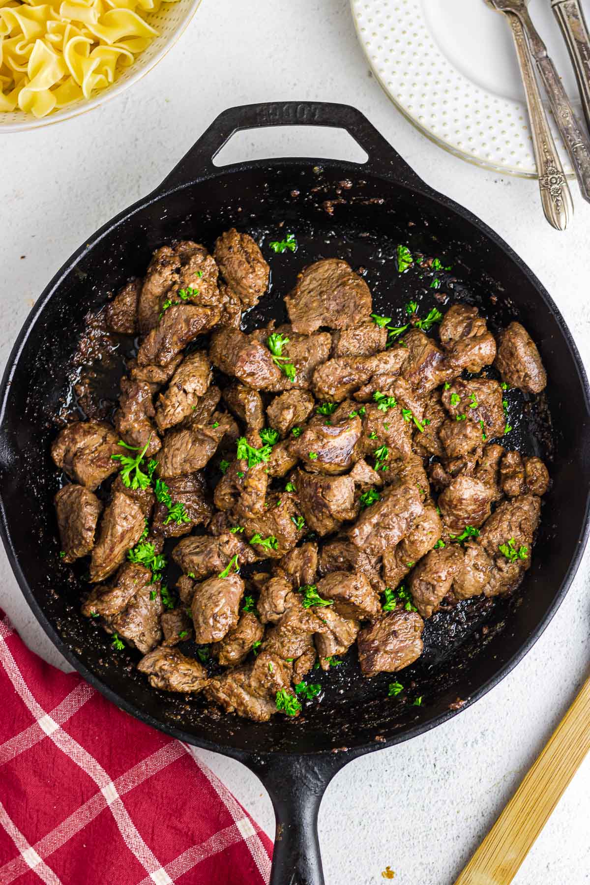 A skillet full of garlic butter steak tips. A bowl of noodles sits off to the side.