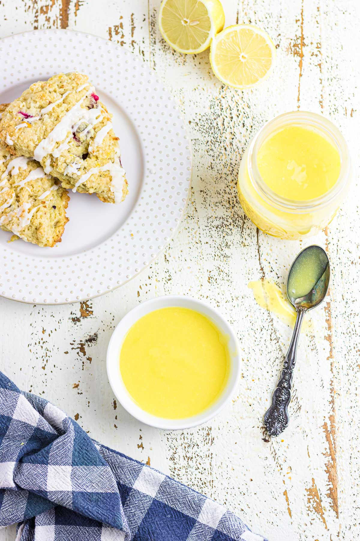 An overhead photo of a bowl and jar filled with lemon curd next to some scones and a spoon coated in curd.