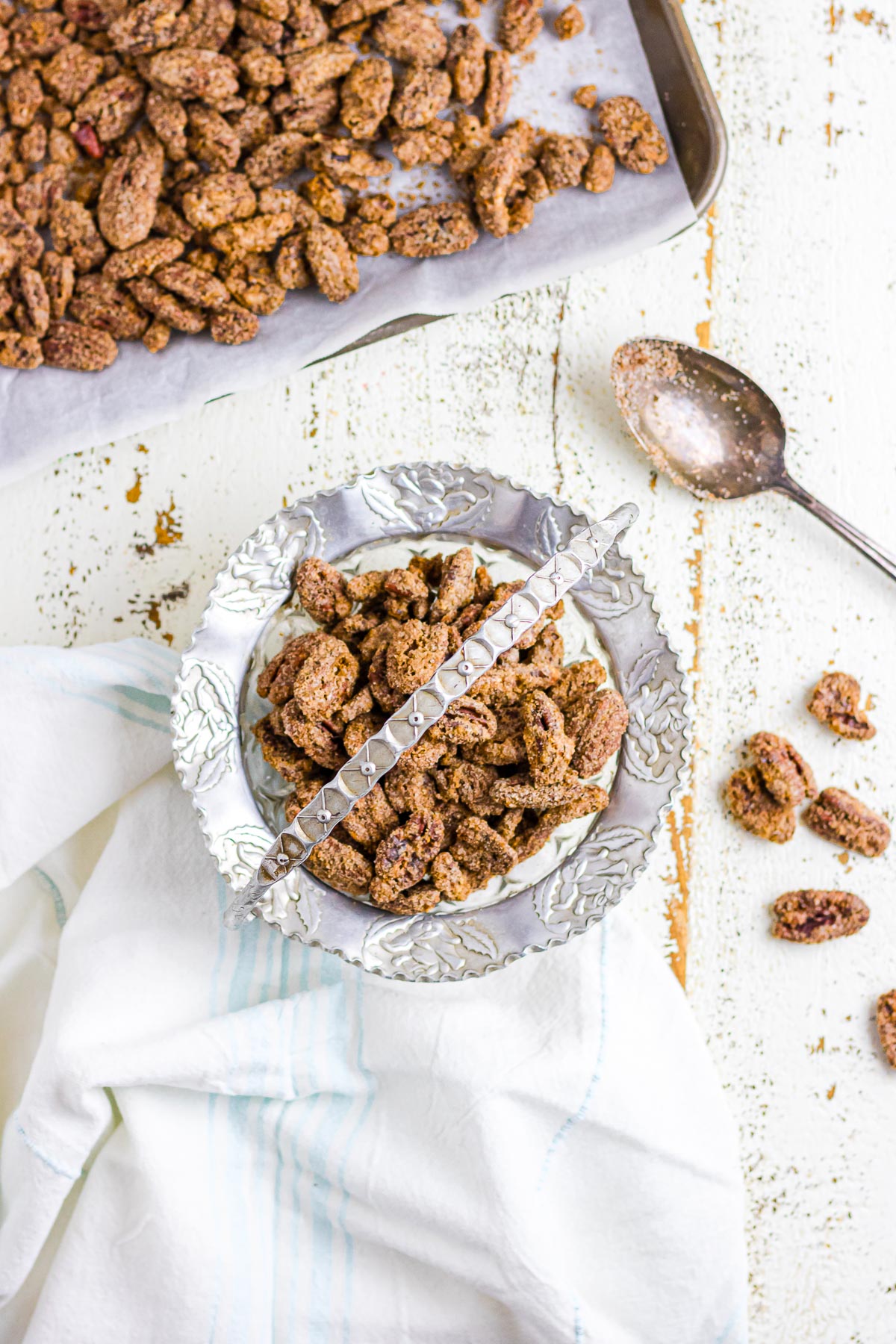 An overhead image of spiced candied pecans in a decorative bowl next to a spoon and baking sheet.
