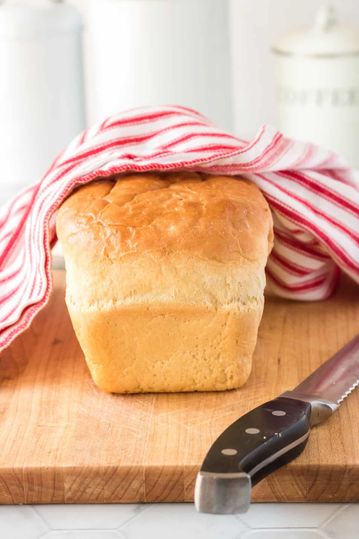 A loaf of batter bread on a cutting board partially covered with a red and white striped tea towel.