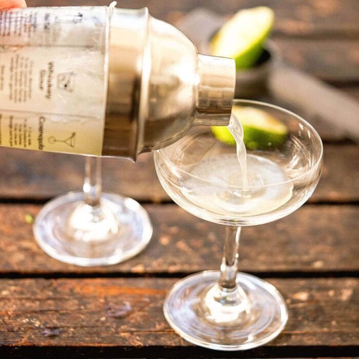 A cocktail shaker pouring the cocktail into a stemmed glass.