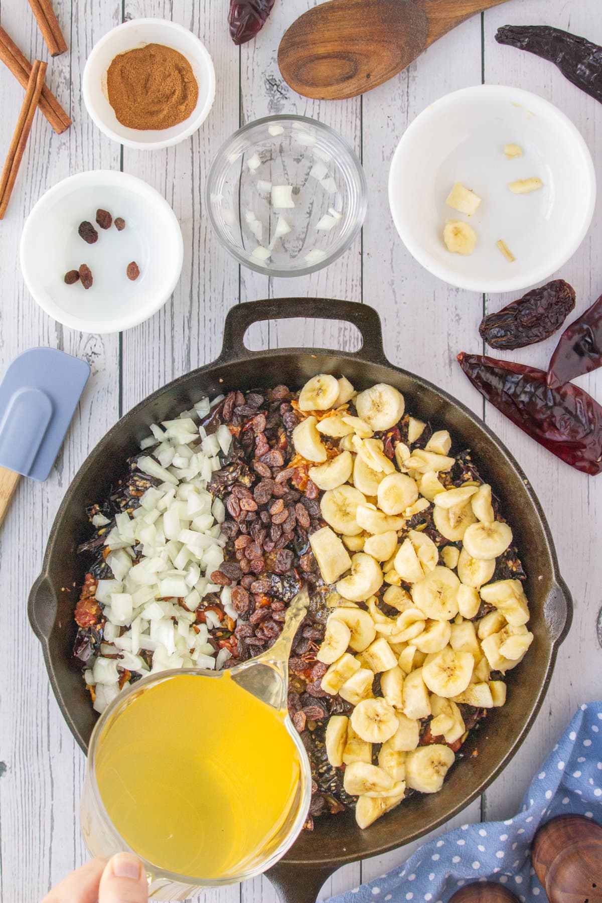 A cup pourig chicken stock into the skillet. bananas, raisins, and onions have been added.
