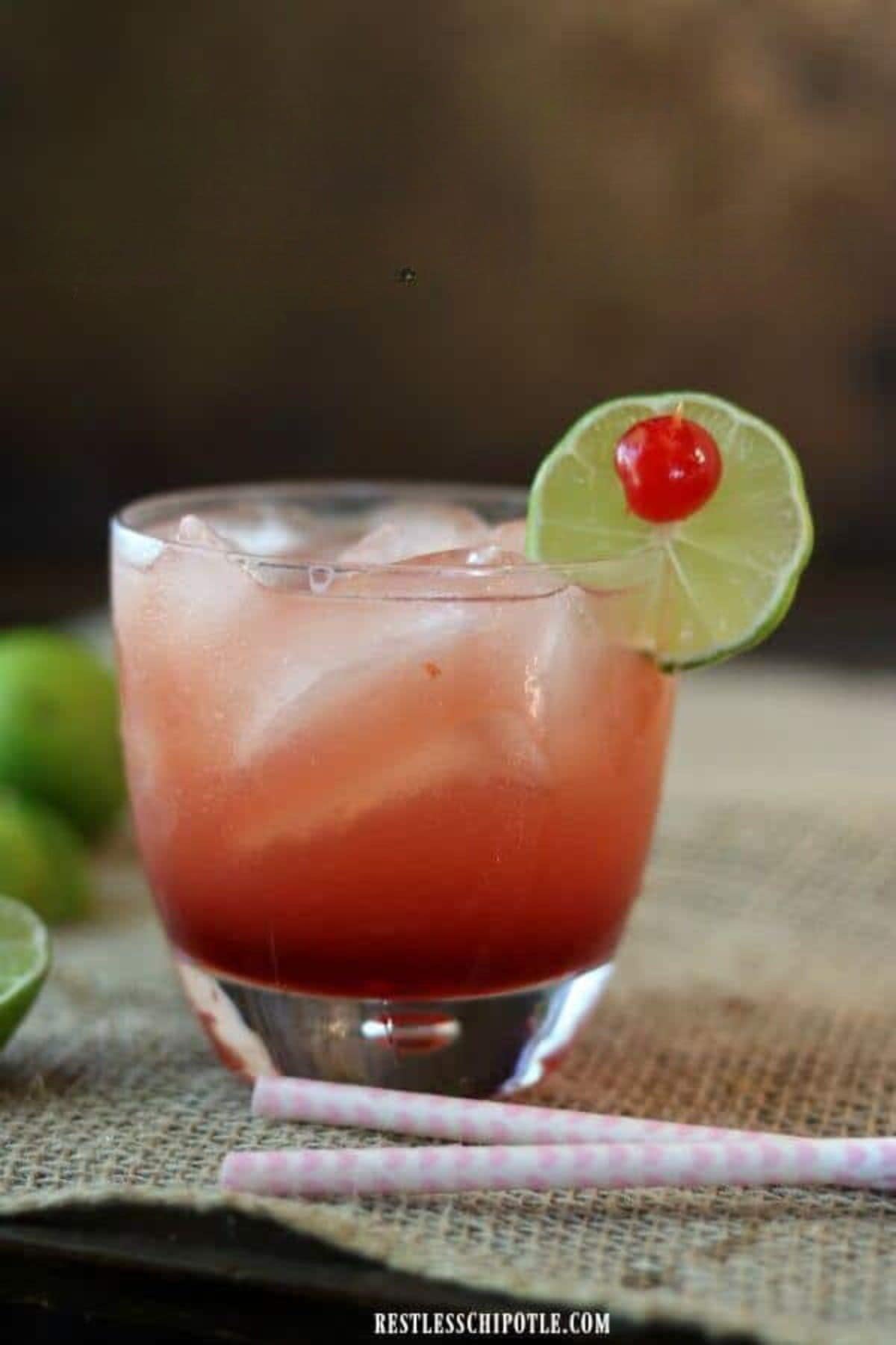 An orange color cocktail in a glass garnished with lime and cherries.