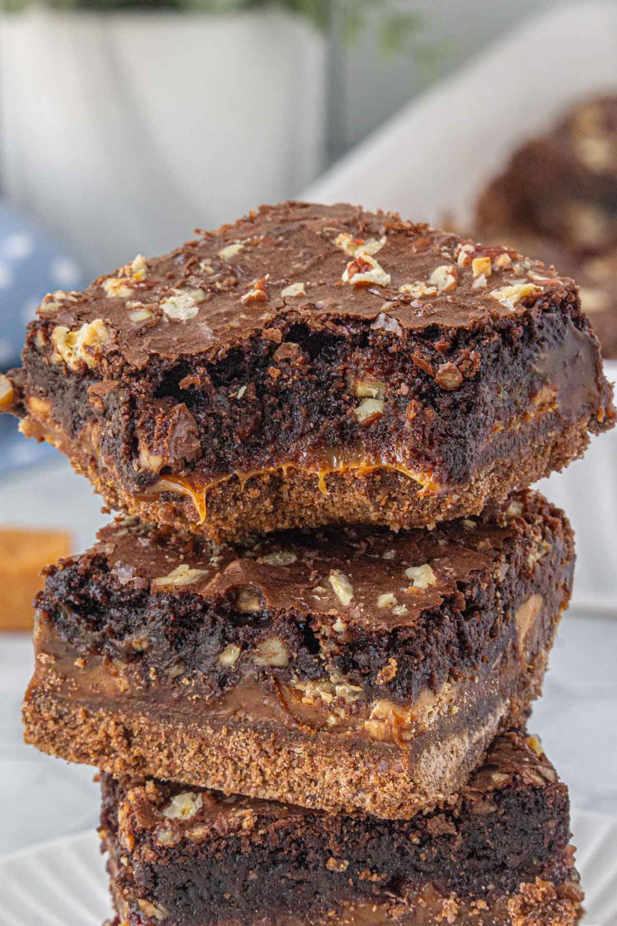 Stack of rich caramel fudge brownies with a graham cracker crust and chopped pecans, showing gooey caramel oozing from the center and a crackly top.