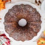 “Overhead view of a homemade apple sour cream bundt cake dusted with powdered sugar, surrounded by fresh apples, pecans, and fall flowers on a rustic white table.”