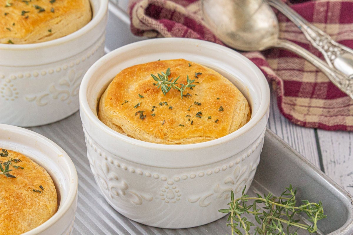 Individual freezer-friendly chicken and gravy casseroles baked in white ramekins with golden biscuit tops, garnished with parsley and fresh thyme.