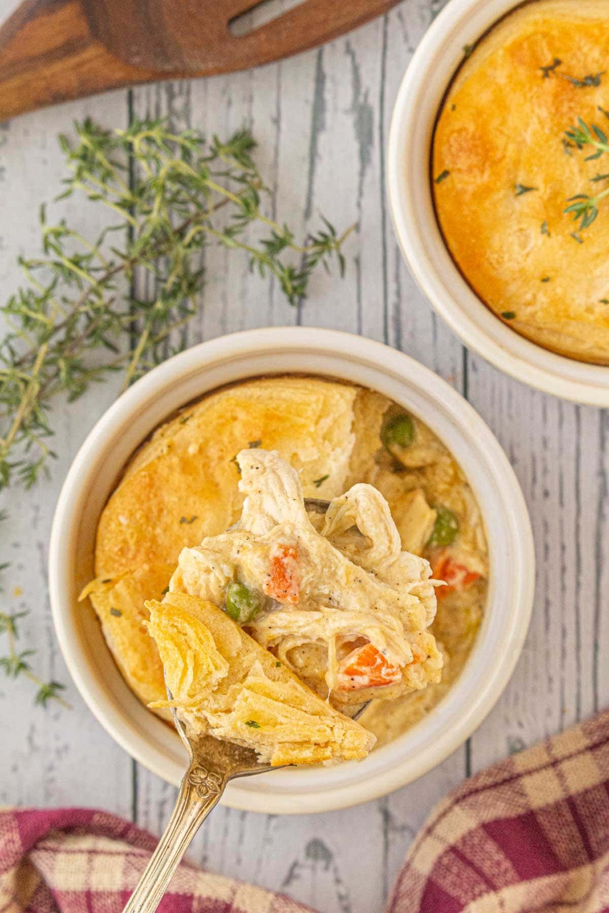 Overhead view of freezer-friendly chicken and gravy casserole in a white ramekin, with a spoon lifting creamy chicken, carrots, peas, and flaky biscuit topping.