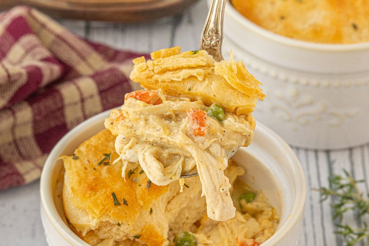 Close-up of a spoonful of creamy chicken and gravy casserole with peas, carrots, and a golden biscuit topping, served in a white ramekin.