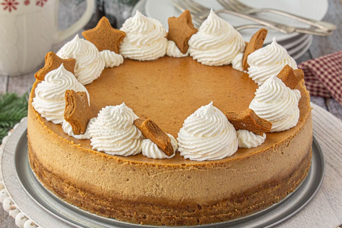 Christmas gingerbread cheesecake topped with whipped cream swirls and gingerbread cookies on a wooden platter. It's a festive holiday cheesecake recipe that's perfect for parties and dessert tables.