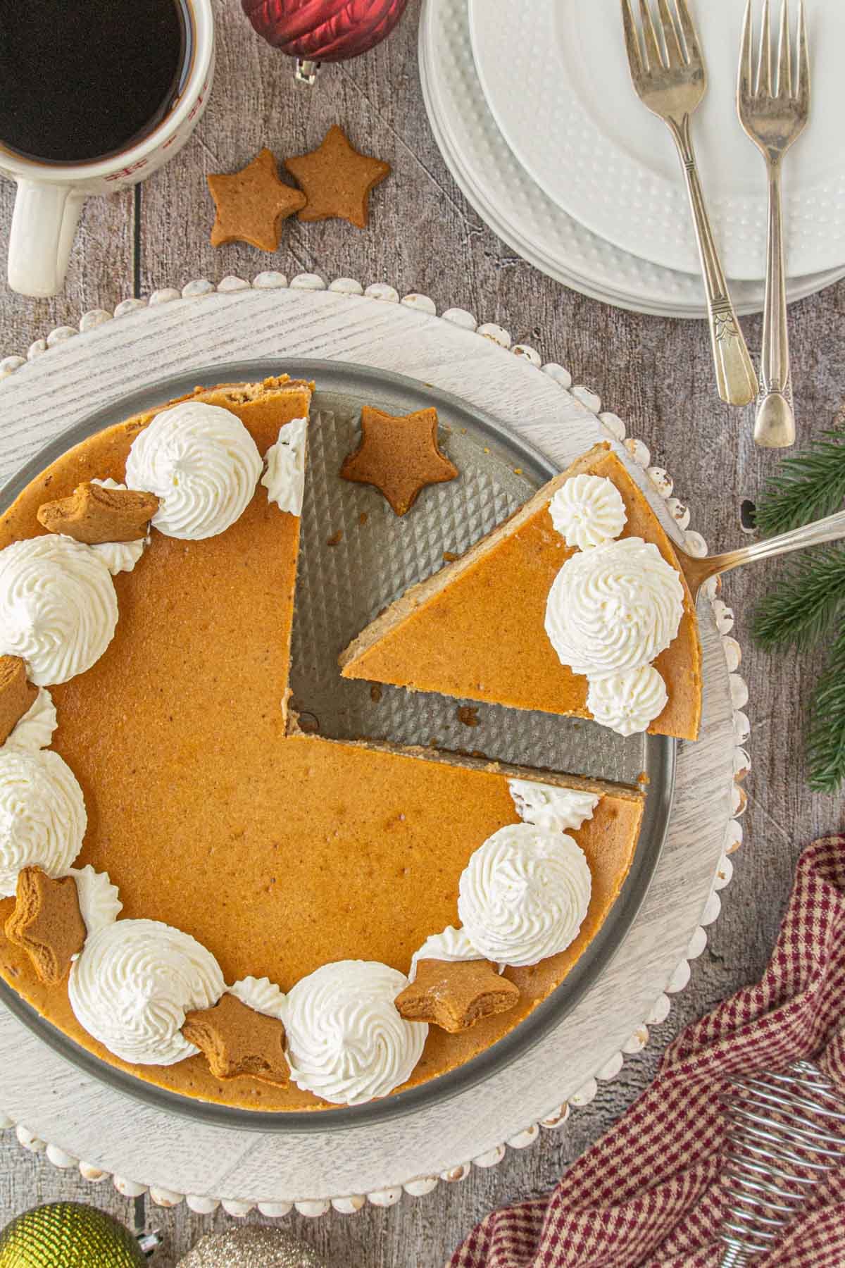 Overhead view of a gingerbread cheesecake with whipped cream swirls and star-shaped cookies, with one slice being lifted from the pan on a rustic holiday table.